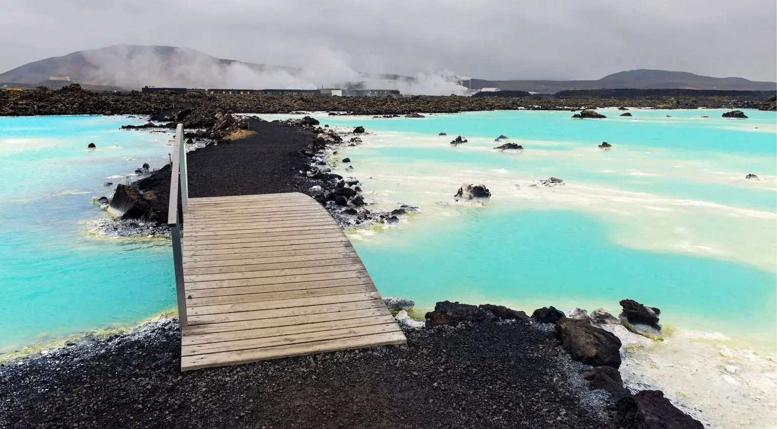 Wooden boardwalk crossing the turquoise Blue Lagoon with the geothermal plant venting steam behind