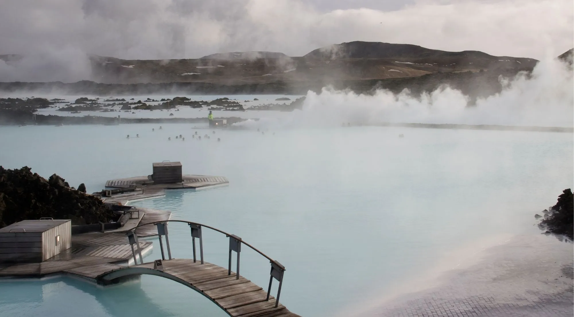 Steam pouring off the Blue Lagoon at dusk with wooden walkways and bathers in the distance