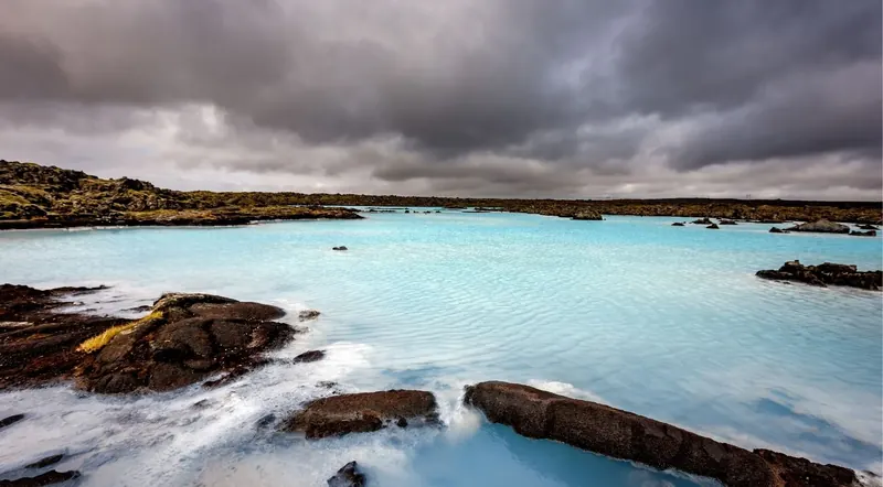 Pale blue geothermal lagoon under a dramatic grey Icelandic sky with lava-rock shoreline