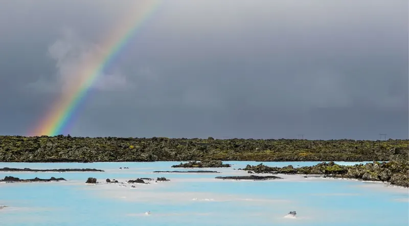 A rainbow arcing over the pale blue silica water and black lava field of the Blue Lagoon