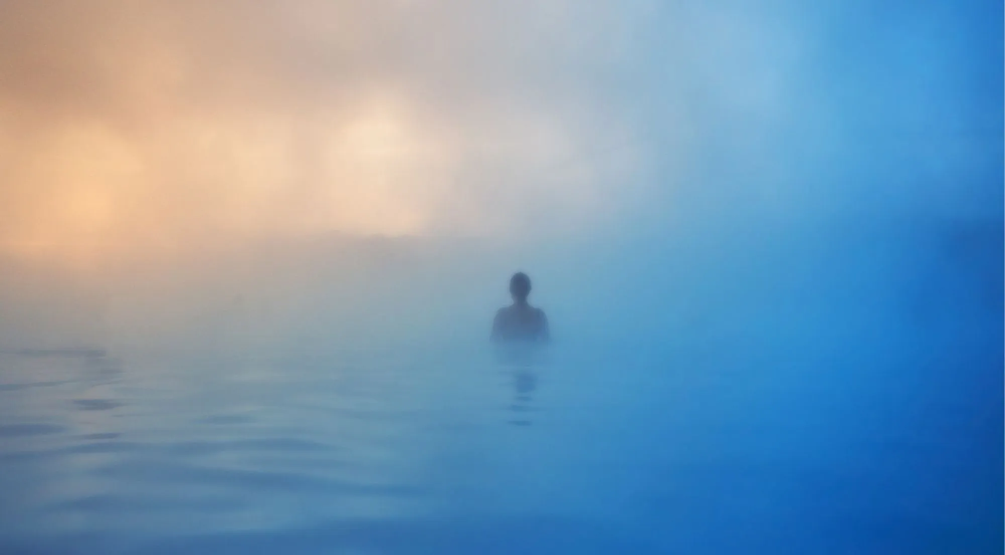 A lone bather silhouetted in thick geothermal steam at dawn, blue water meeting golden sky