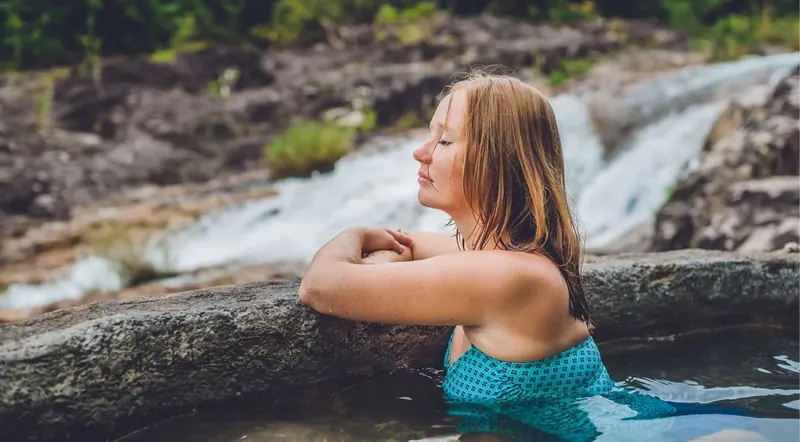 A woman soaking in a natural stone hot spring with a waterfall cascading behind her