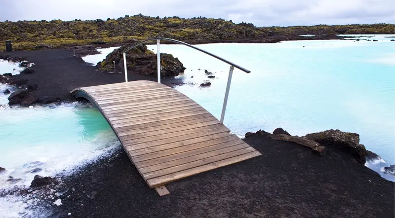 Bright turquoise silica water lapping against black volcanic rocks under a moody sky